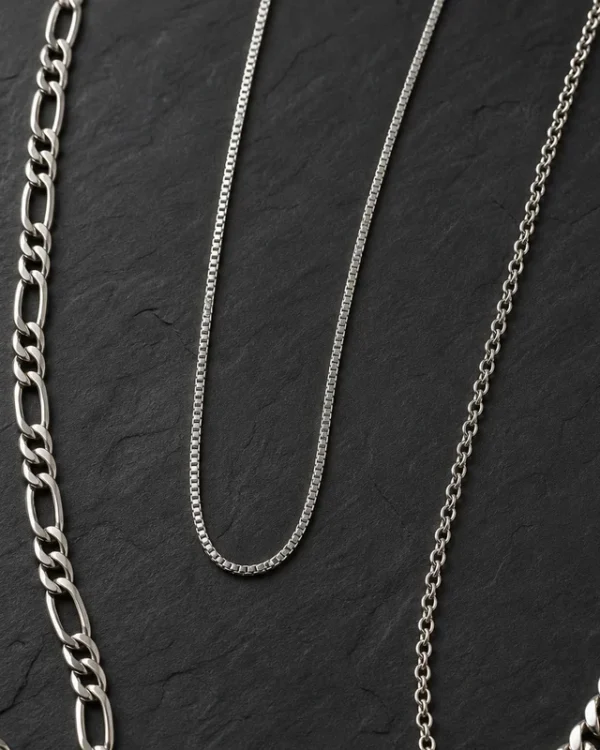 Plusieurs chaînes et colliers homme en argent et acier posés sur une surface sombre, différentes mailles et longueurs